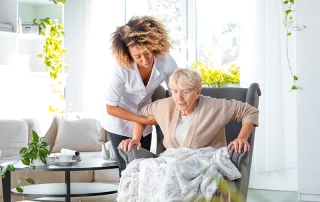 Home carer giving support of old woman to get up from an armchair in her room